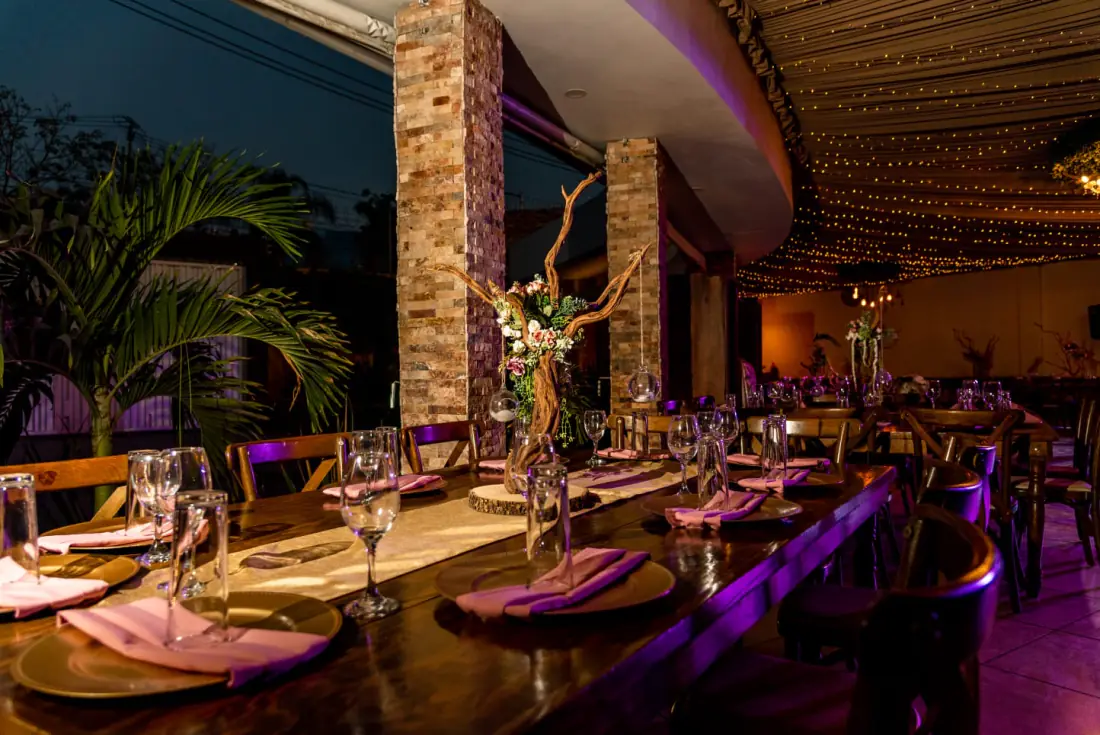 Terraza de noche: mesa larga de madera con servilletas rosas bajo el cielo de luces cálidas, junto a la palmera